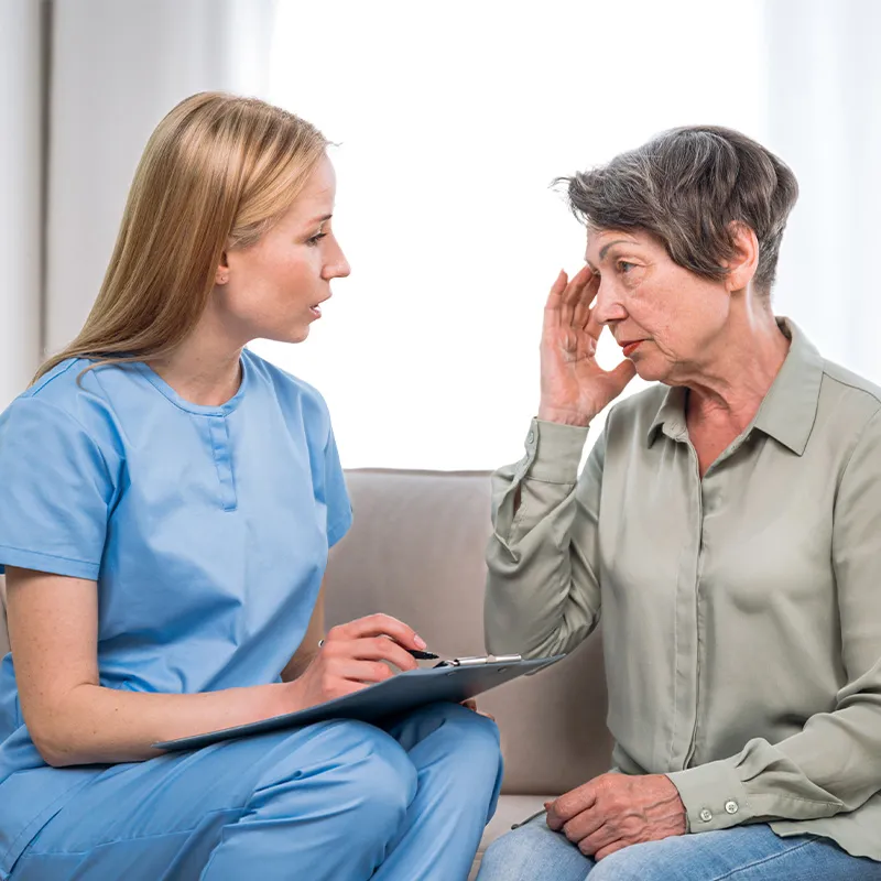 A clinician in scrubs sitting with a notepad, listening to an older woman on a couch who is resting a hand against her temple.