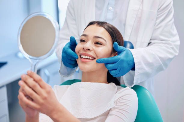 A patient in a dental chair smiling into a hand mirror while a dentist in blue gloves checks her teeth.