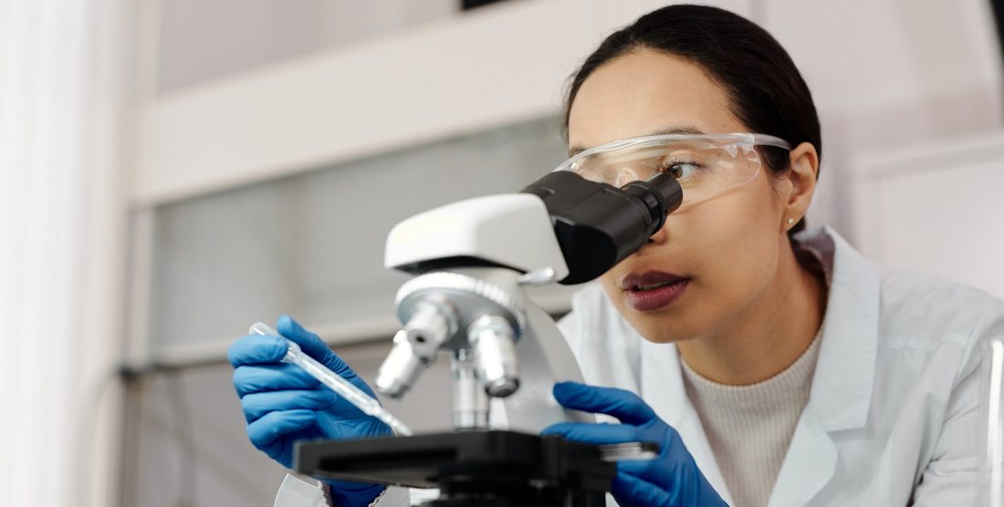A laboratory scientist in a white coat and safety glasses examining a sample under a microscope.
