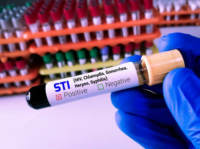 A gloved hand holding a blood sample tube labelled as an STI panel for HIV, chlamydia, gonorrhea, herpes, and syphilis, in front of racks of collection tubes.